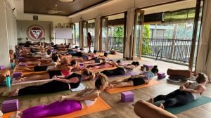 People practicing yoga on mats in a spacious room with open windows; yoga props are placed around them.