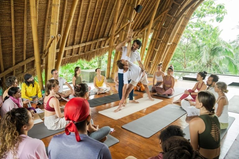 Yoga instructor assists a participant with a pose in a bamboo pavilion, surrounded by a seated group on mats, amidst greenery.