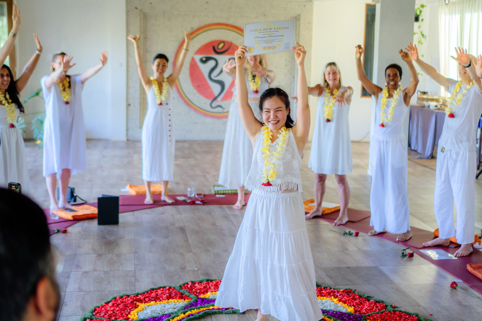 A person in white holding a certificate, surrounded by others in a circle, all wearing leis. A colorful flower design is on the floor.