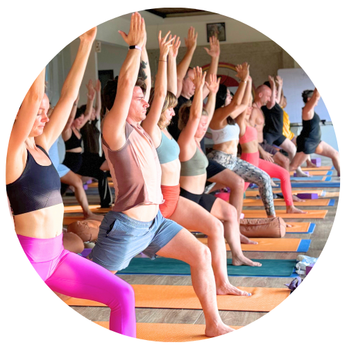 A group of people practicing yoga indoors, performing a standing pose with raised arms. They are on orange and blue mats, facing forward.
