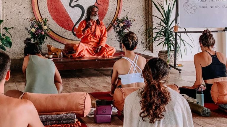 A person in an orange robe sitting cross-legged on a platform, speaking to a group in yoga pose. Plants and a circular mural in the background.