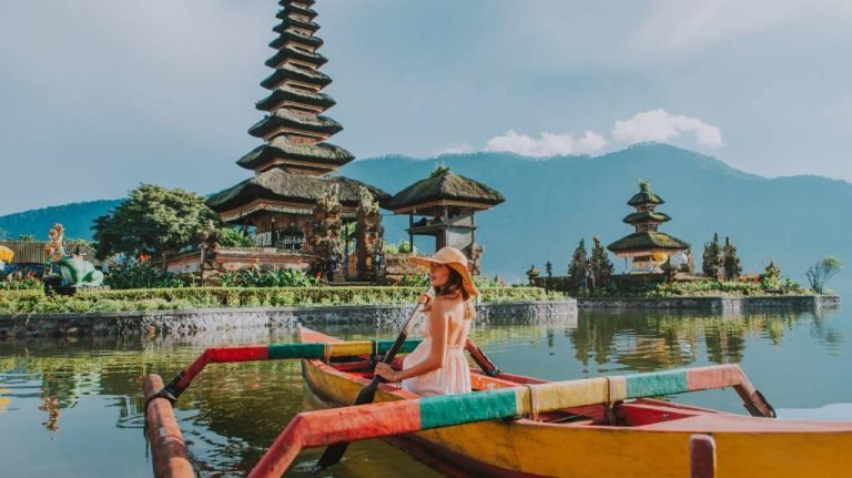 A person in a straw hat sits in a colorful boat on a lake, with a multi-tiered pagoda and lush greenery in the background.