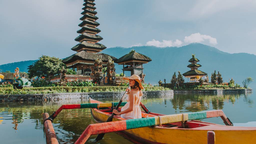 A person in a straw hat sits in a colorful boat on a lake, with a multi-tiered pagoda and lush greenery in the background.