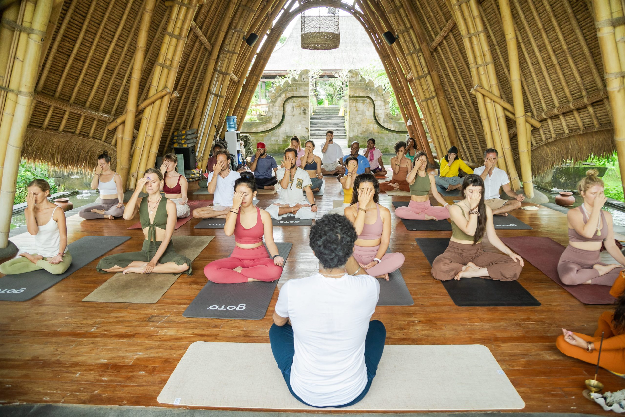 People in a bamboo structure practicing yoga and meditation, sitting cross-legged on mats, with greenery visible outside.