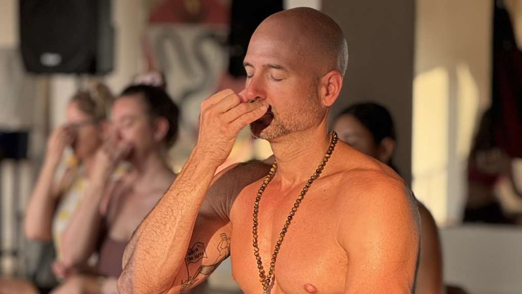 A man practicing breathing exercises in a yoga class, eyes closed, wearing a beaded necklace. Others in the background doing the same.