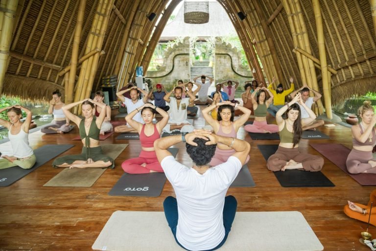 A group of people practicing yoga inside a bamboo structure. They are seated on mats, hands on heads, with a large entrance visible ahead.