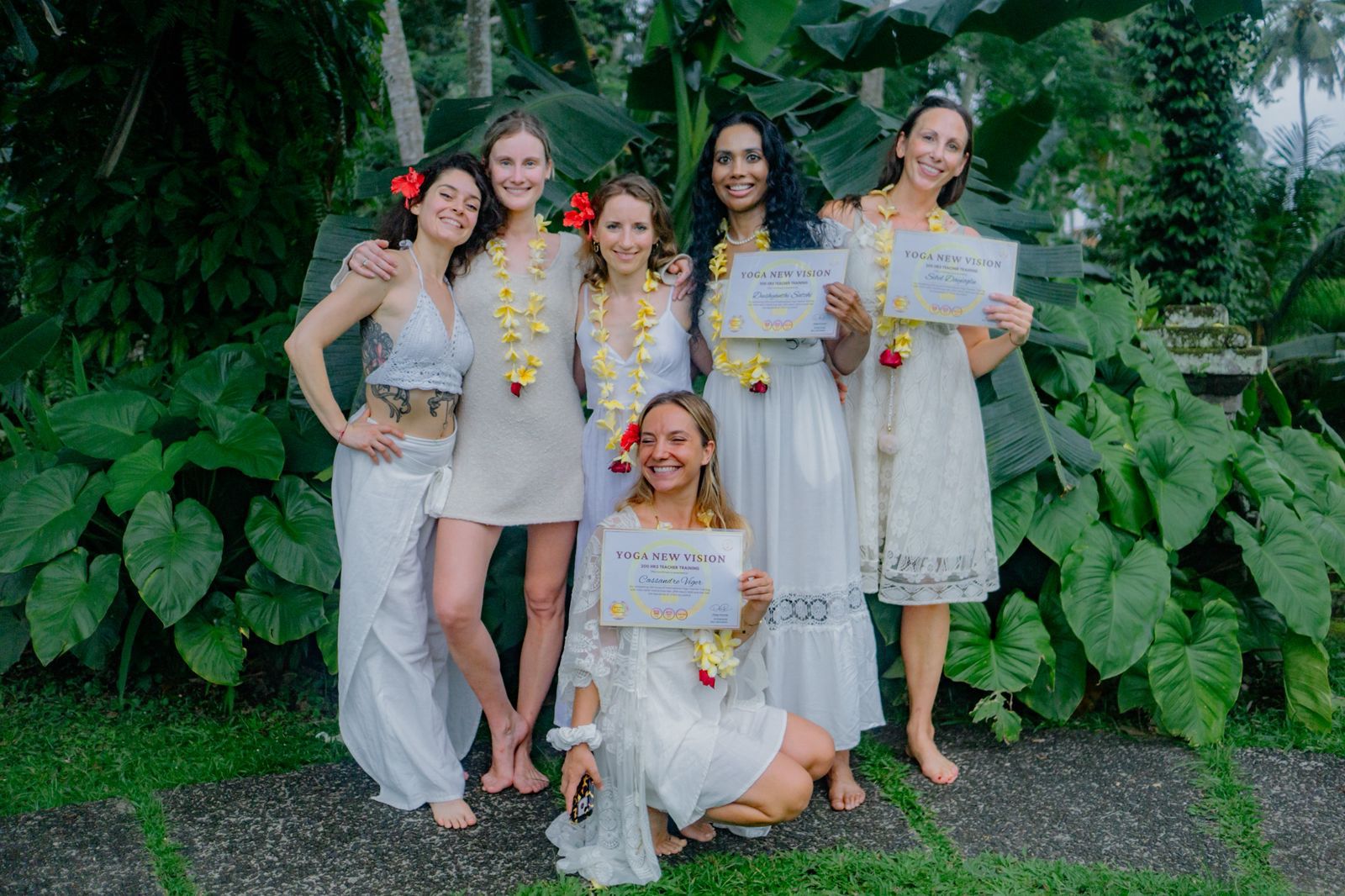 Six women in white outfits hold certificates and wear flower garlands, standing and kneeling in front of lush green plants outdoors.